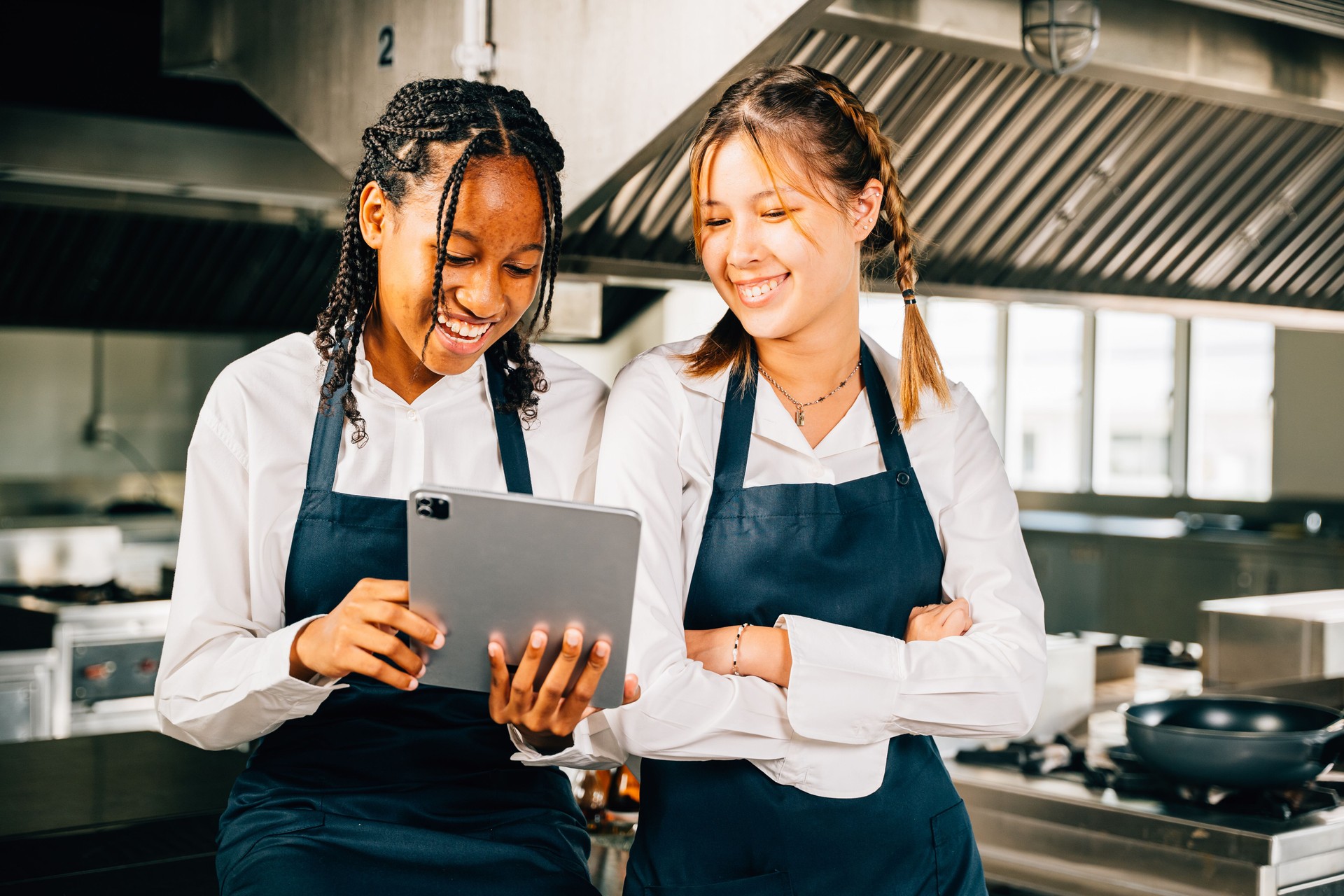 Two famous chefs in stainless steel kitchen discuss video blog using tablet. Professionals smiling standing reading searching. Modern technology in restaurant industry holding device.